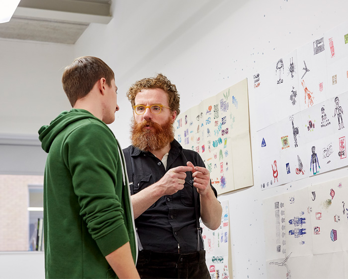 A student and a tutor stood talking next to illustrations on a wall