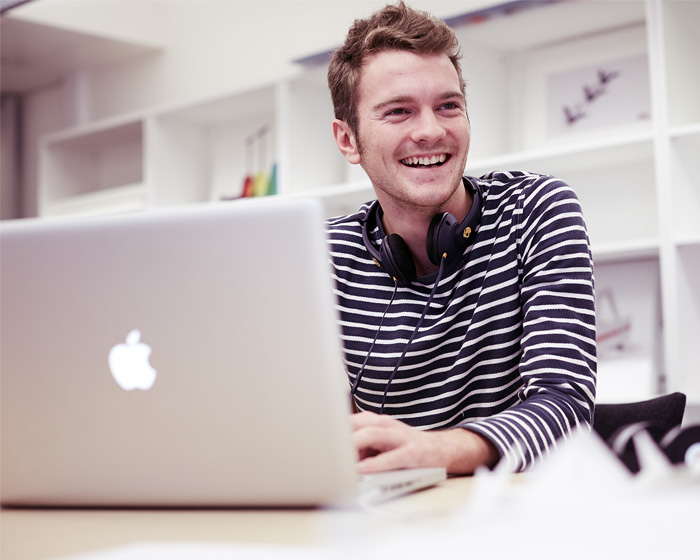 A student sat with a Macbook and headphones smiling off camera