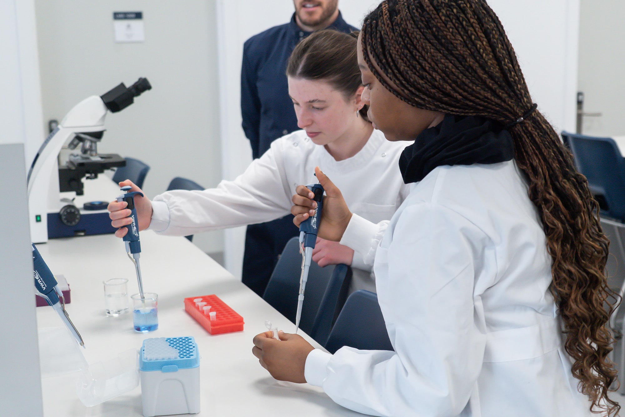 Two students in a lab wearing white lab coats dropping liquid into containers, whilst a lecturer observes